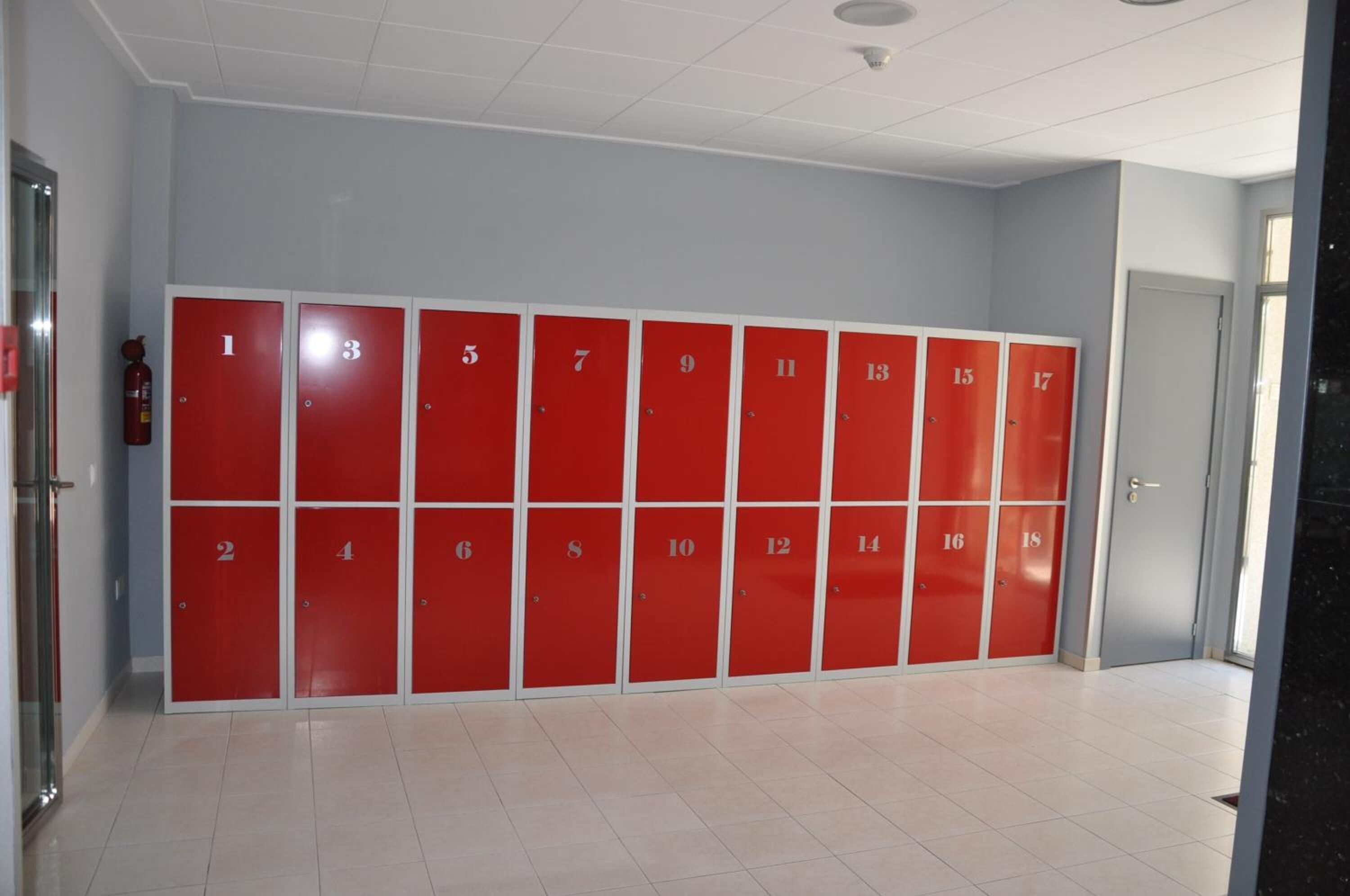 A row of red lockers numbered 1 to 18 in a hallway with white tiled floors and light blue walls.