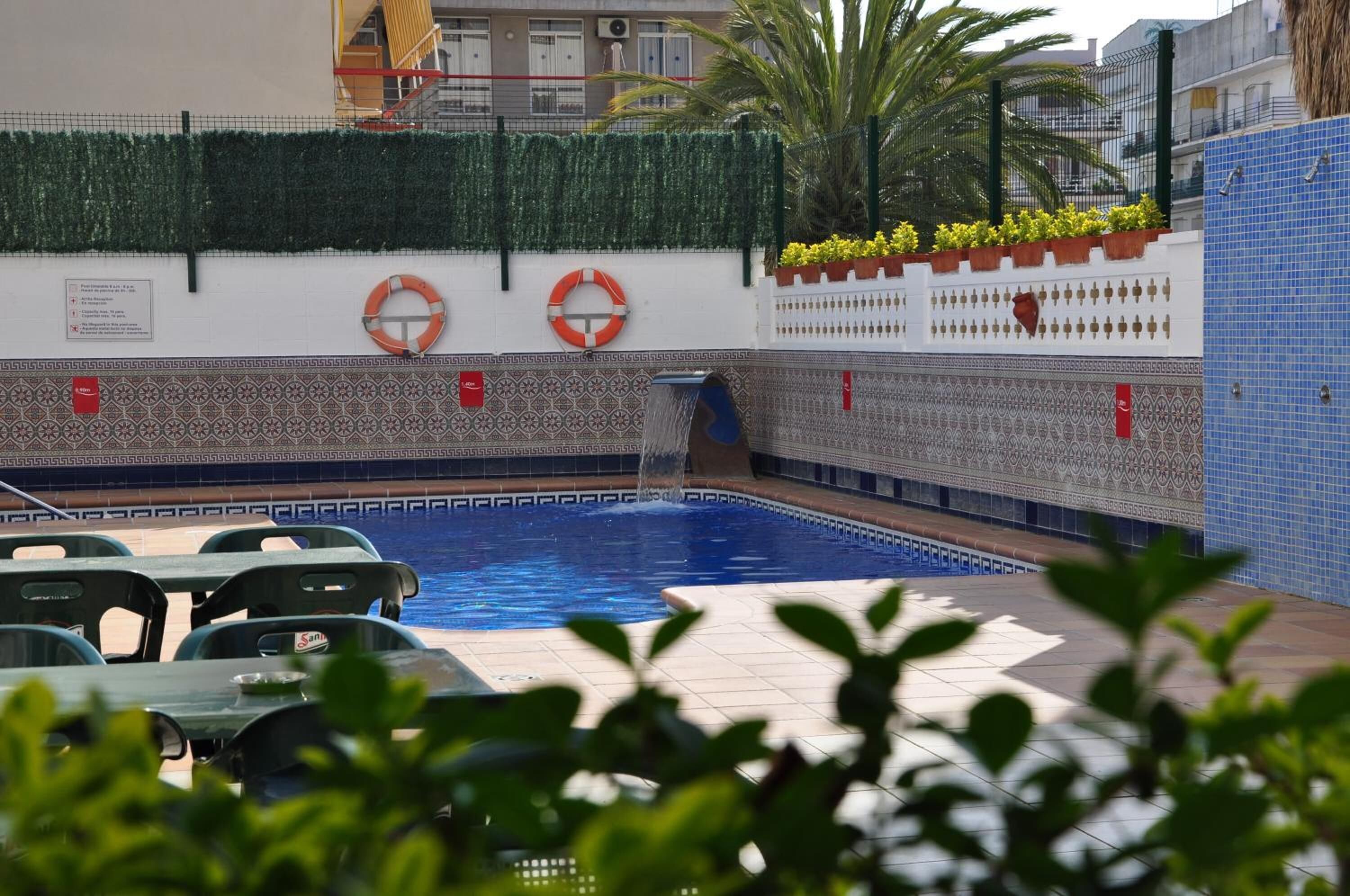 En pool med en vattenfontän, omgiven av en terrass med kakelplattor och stolar.