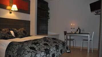Habitación de hotel con cama, lámpara de noche, televisor, escritorio con dos sillas y una mesita con velas y copas.