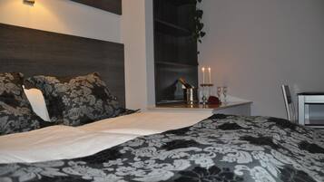 Une chambre à coucher avec un grand lit, une table de chevet sur laquelle se trouvent une bouteille et des bougies, et une fenêtre avec une plante.