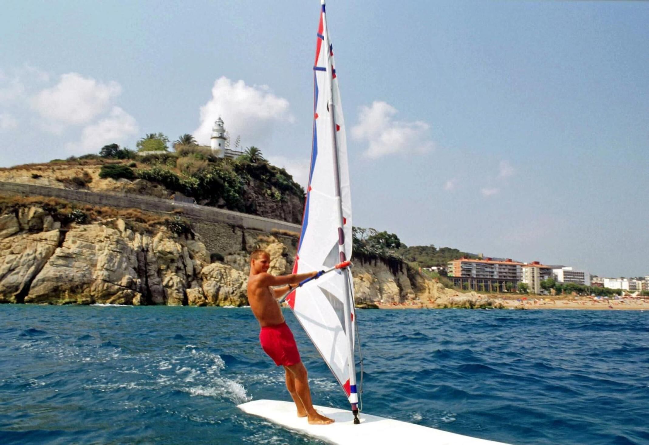 Una persona practicando windsurf en un día despejado, con un litoral rocoso y edificios al fondo.