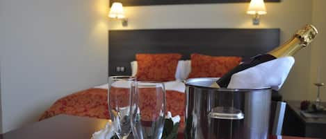 Habitación de hotel con una cama, dos lámparas de noche, un minibar con una botella de champán y dos copas sobre una mesa.