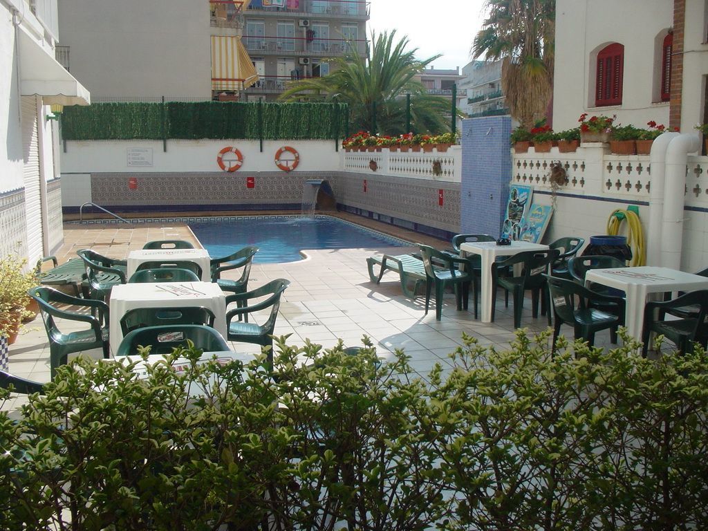 An outdoor pool area with green chairs and tables, surrounded by a white wall with red accents and potted plants.