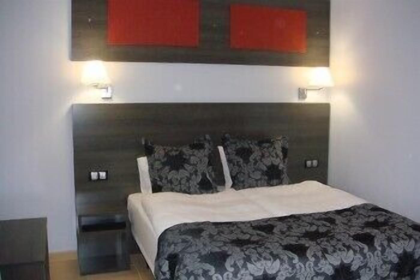 A bedroom with a dark headboard, two bedside lamps, a bed with a patterned bedspread, and a small nightstand.