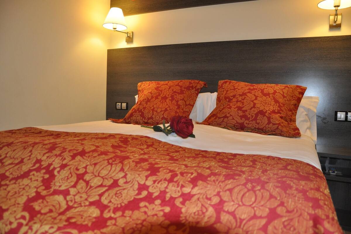 A hotel room with a large bed, two red patterned pillows, a single red rose, and two wall-mounted lamps.
