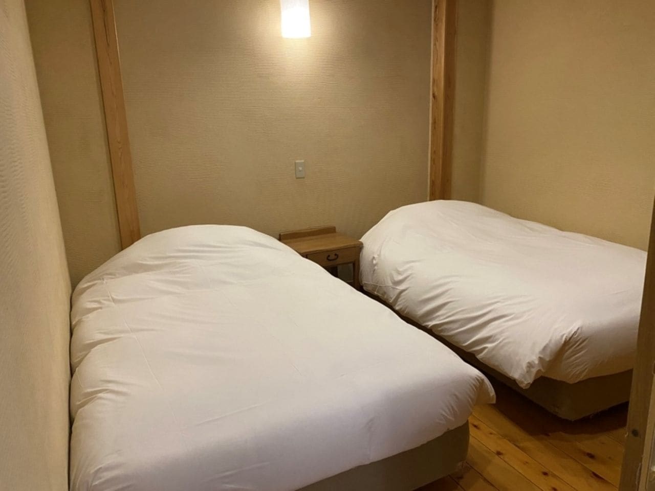 A small, compact room with two single beds, a nightstand, and a wall-mounted light fixture.