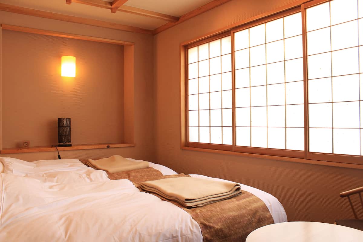 A hotel room with two beds, a large window, a wall-mounted lamp, and a small table.