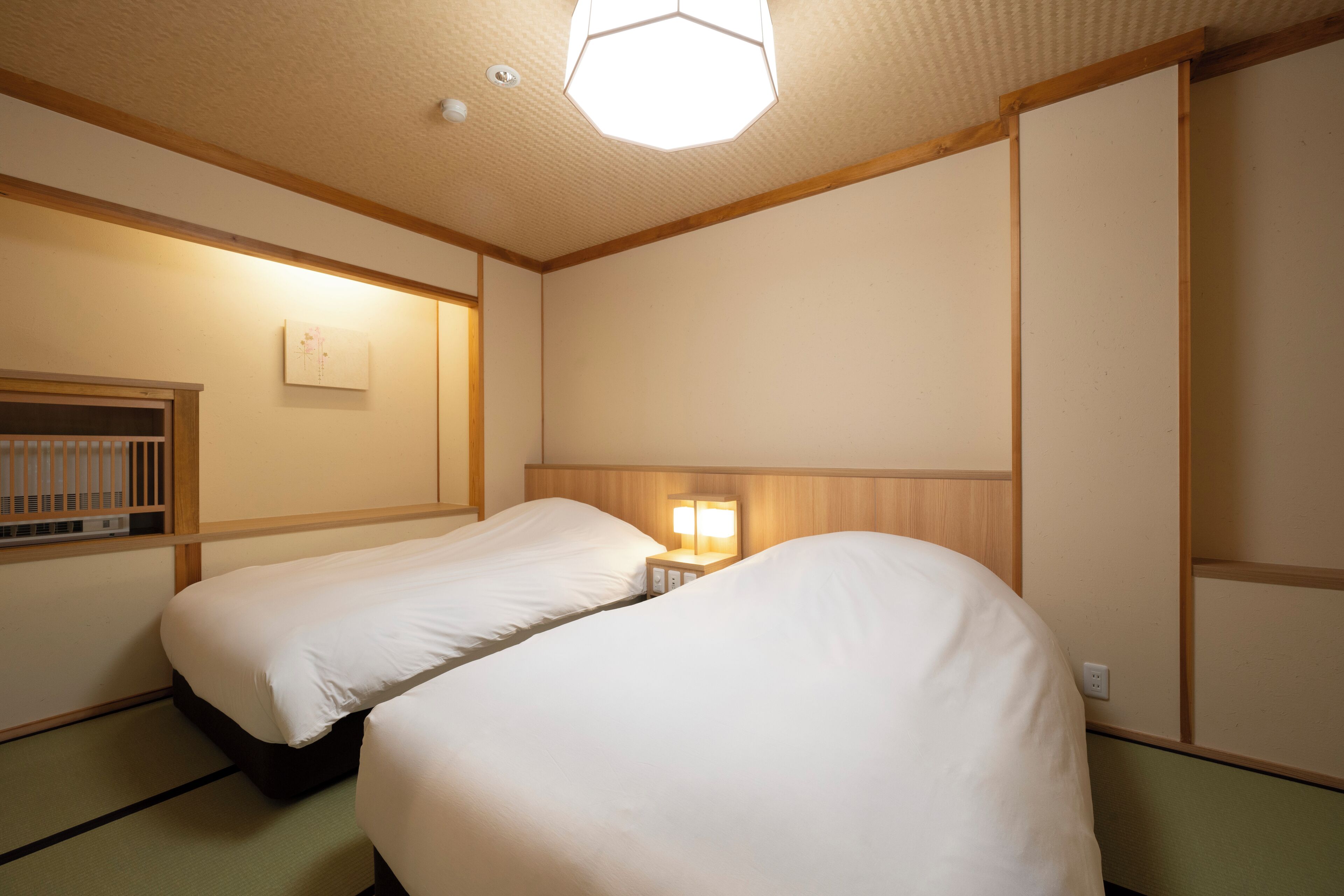 Ein Hotelzimmer mit zwei Einzelbetten, einem Nachttisch mit Lampe, einem kleinen Regal und einem Fenster mit Blick nach draußen.