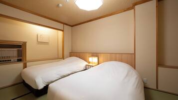 Ein Hotelzimmer mit zwei Einzelbetten, einem Nachttisch mit Lampe, einem kleinen Regal und einem Fenster mit Blick nach draußen.