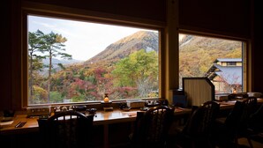 Una habitación con un ventanal que ofrece vistas a un paisaje montañoso con follaje otoñal. En el interior, hay un escritorio de madera con una lámpara, una silla y varios objetos sobre el mismo.