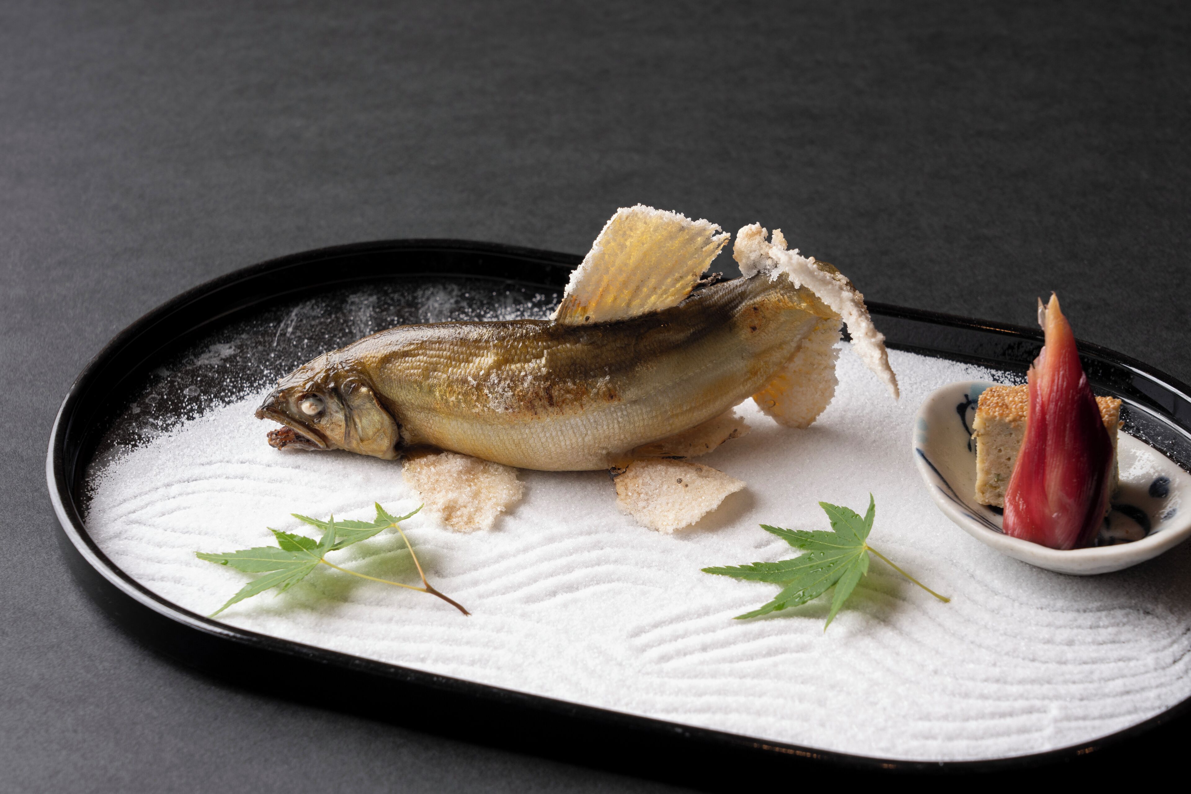 A dish with a fish, garnished with leaves and sugar, on a textured plate.