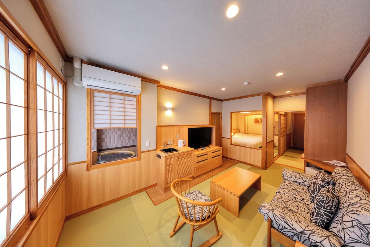 A traditional Japanese-style room with tatami flooring, a low wooden table, a sofa with patterned cushions, a television, and a sliding door leading to a bedroom.