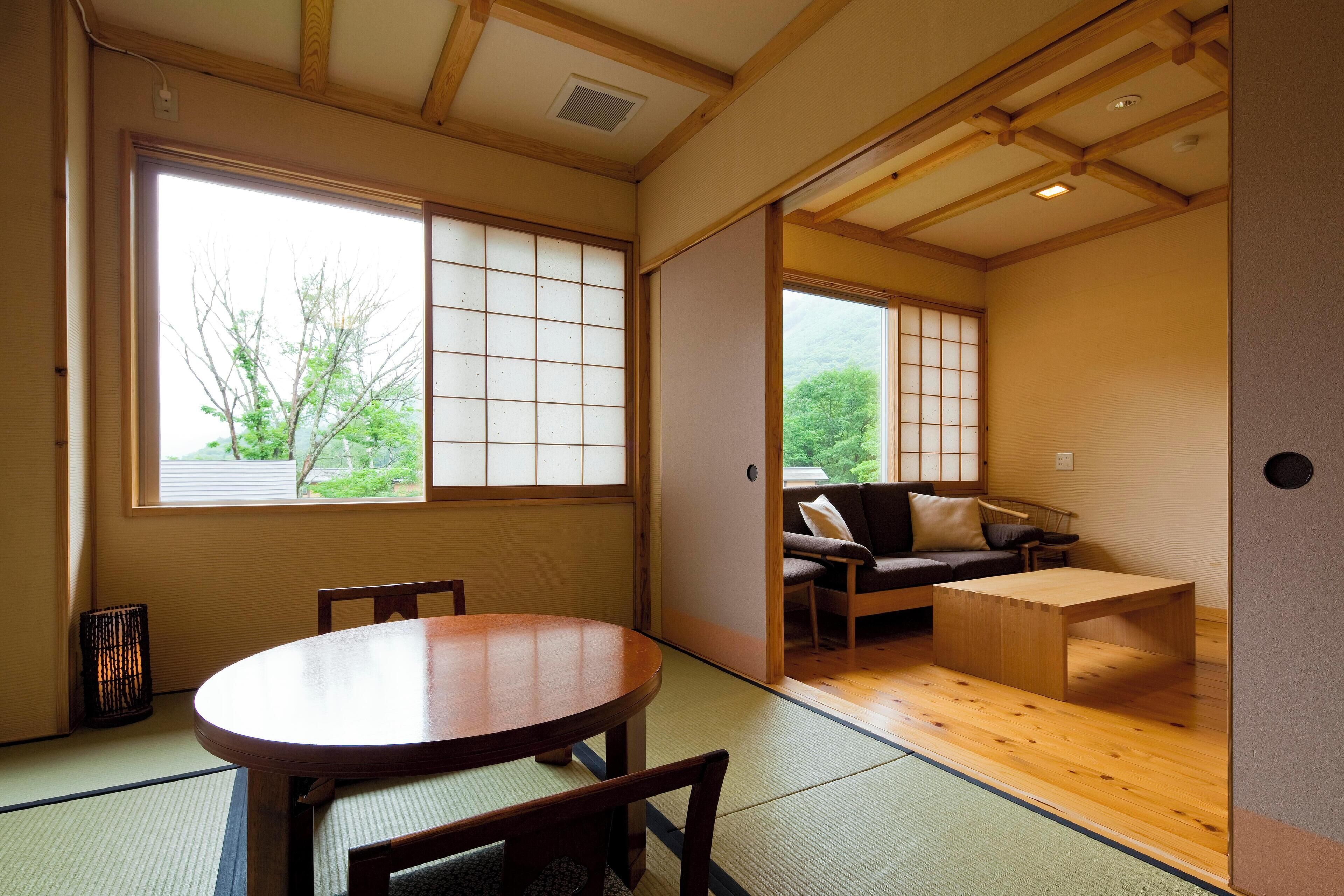 Una habitación japonesa tradicional con suelo de tatami, una mesa de madera baja y una puerta corredera que da a una sala de estar con un sofá y una pequeña mesa.