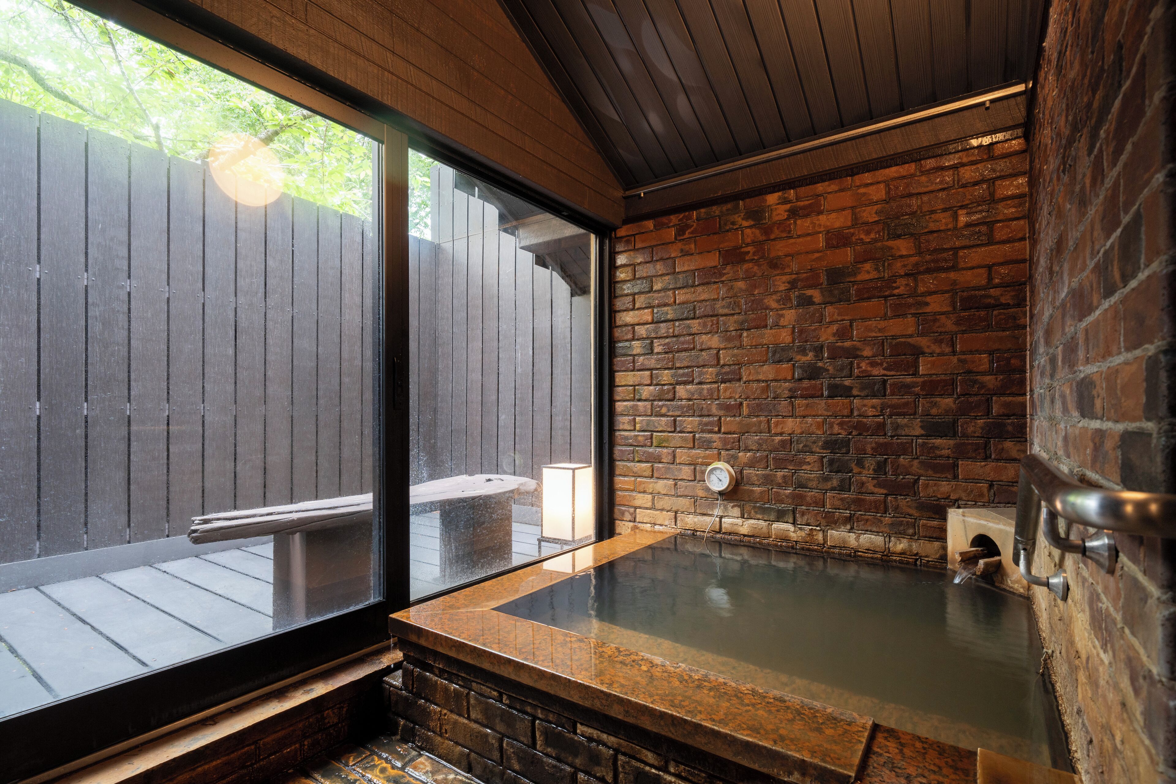 Un espacio de spa con cerramiento de vidrio, pared de ladrillo y una terraza de madera visible en el exterior.