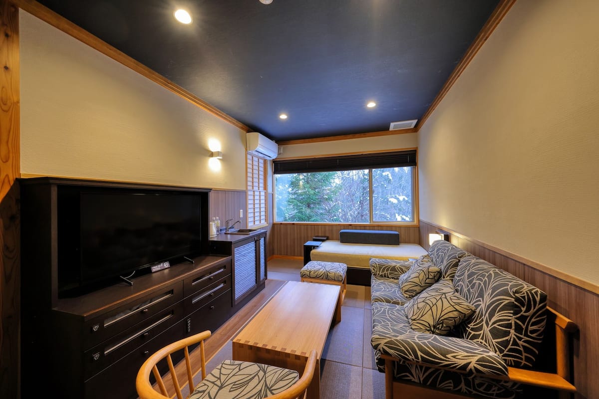 A modern living room with a flat-screen TV, wooden furniture, and a large window offering a view of trees.