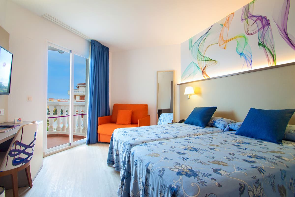 Ein Hotelzimmer mit zwei Betten, einem Schreibtisch und einem Sessel. Es gibt einen Balkon mit Blick auf ein Gebäude.