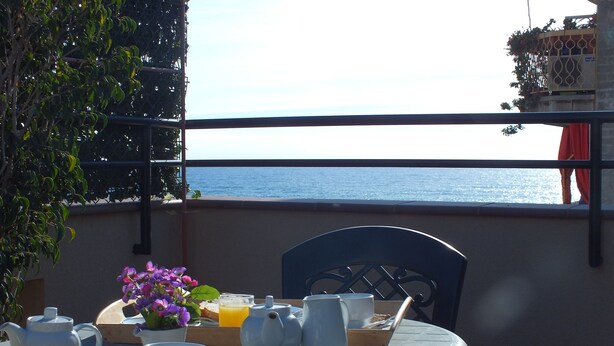 Ein Frühstückstisch mit Tablett, Teekanne, Tassen und Blumen auf einem Balkon mit Meerblick.
