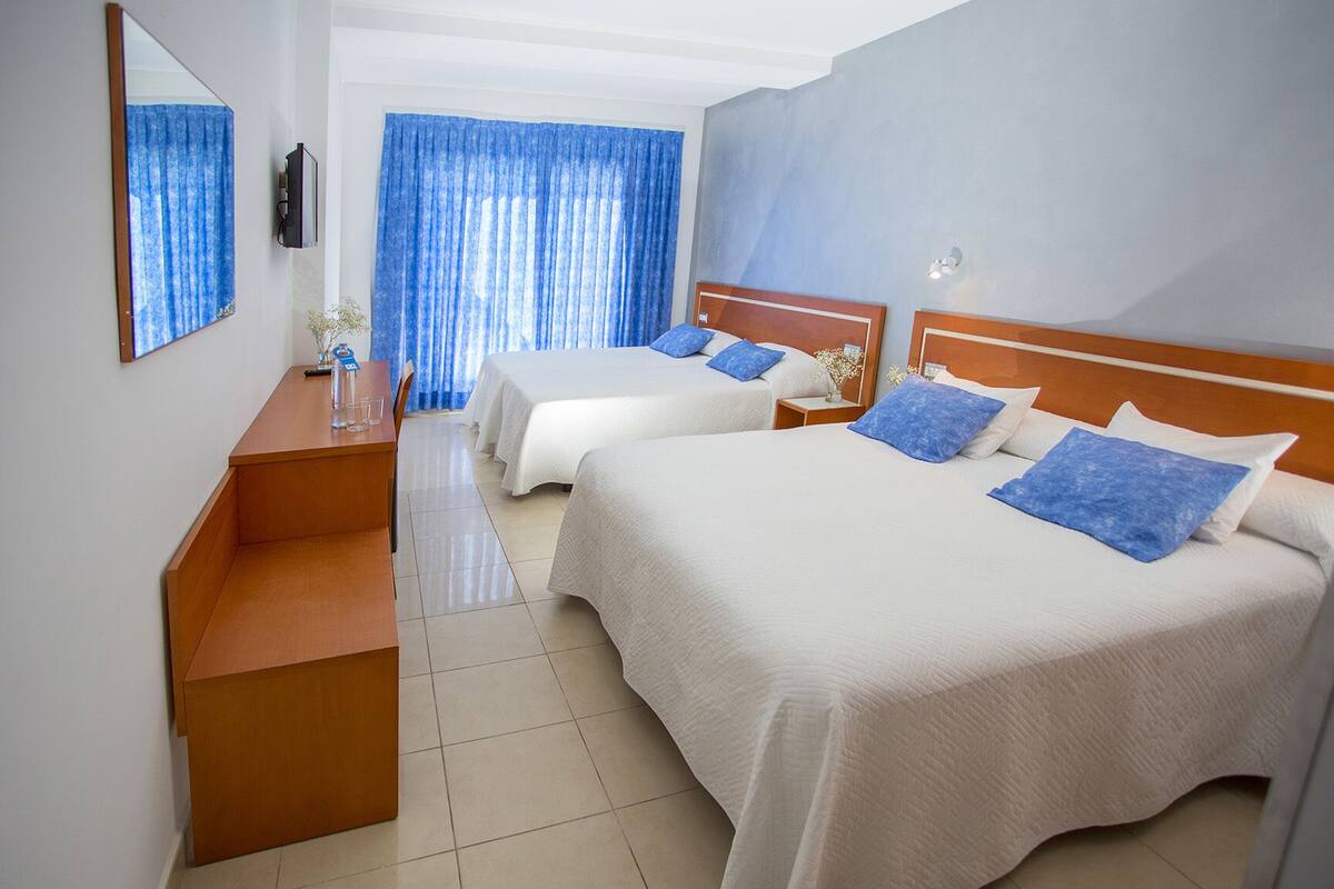 A hotel room with two beds, a wooden desk, a mirror, and a view of the outside through blue curtains.