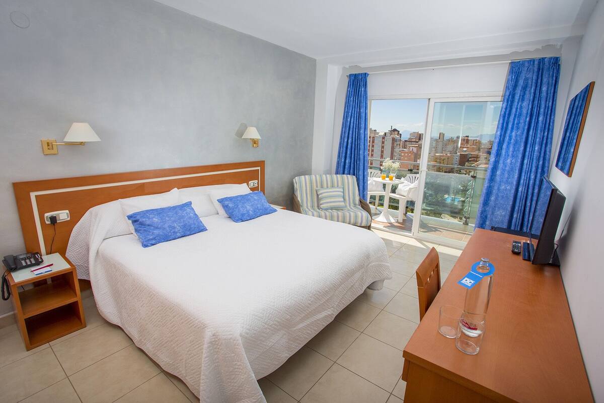 A hotel room with a large bed, a desk with a computer, a chair, a small table with a bottle, and a view of the city through a large window.