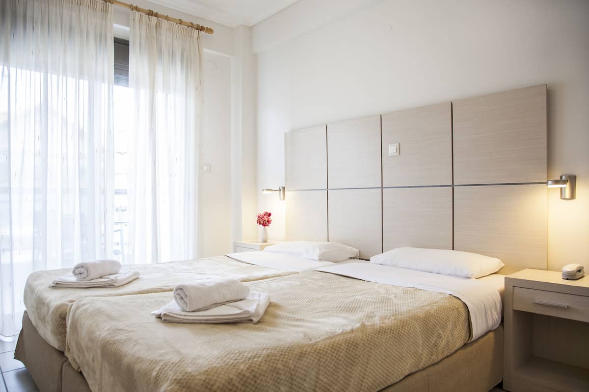 A hotel room with two beds, a bedside table with a lamp, and a window with sheer curtains.