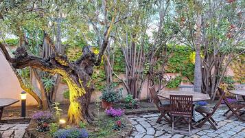 Un patio al aire libre con suelo de piedra, mesas y sillas de madera, y diversas plantas y flores.