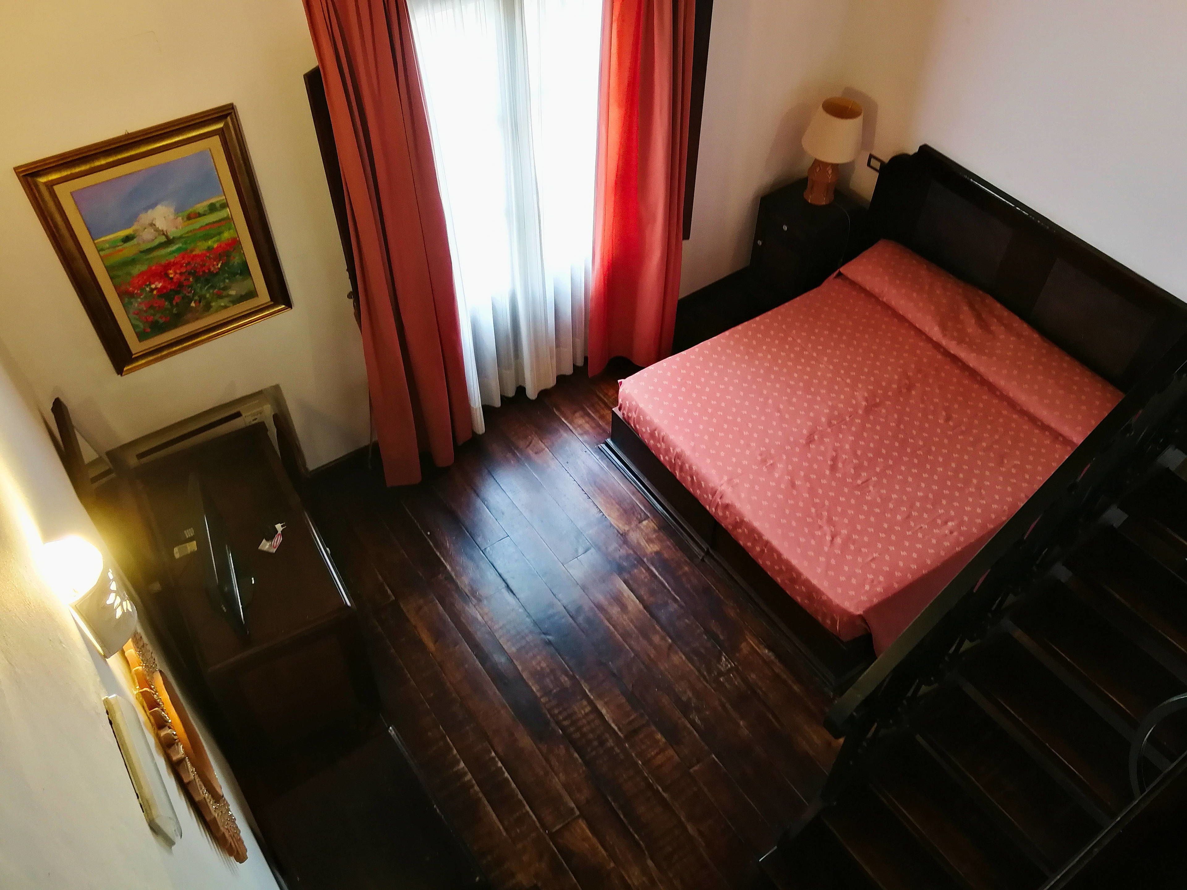 Una habitación con piso de madera, una cama con cubrecama rojo, una mesita de noche con lámpara, un cuadro en la pared y una ventana con cortinas.