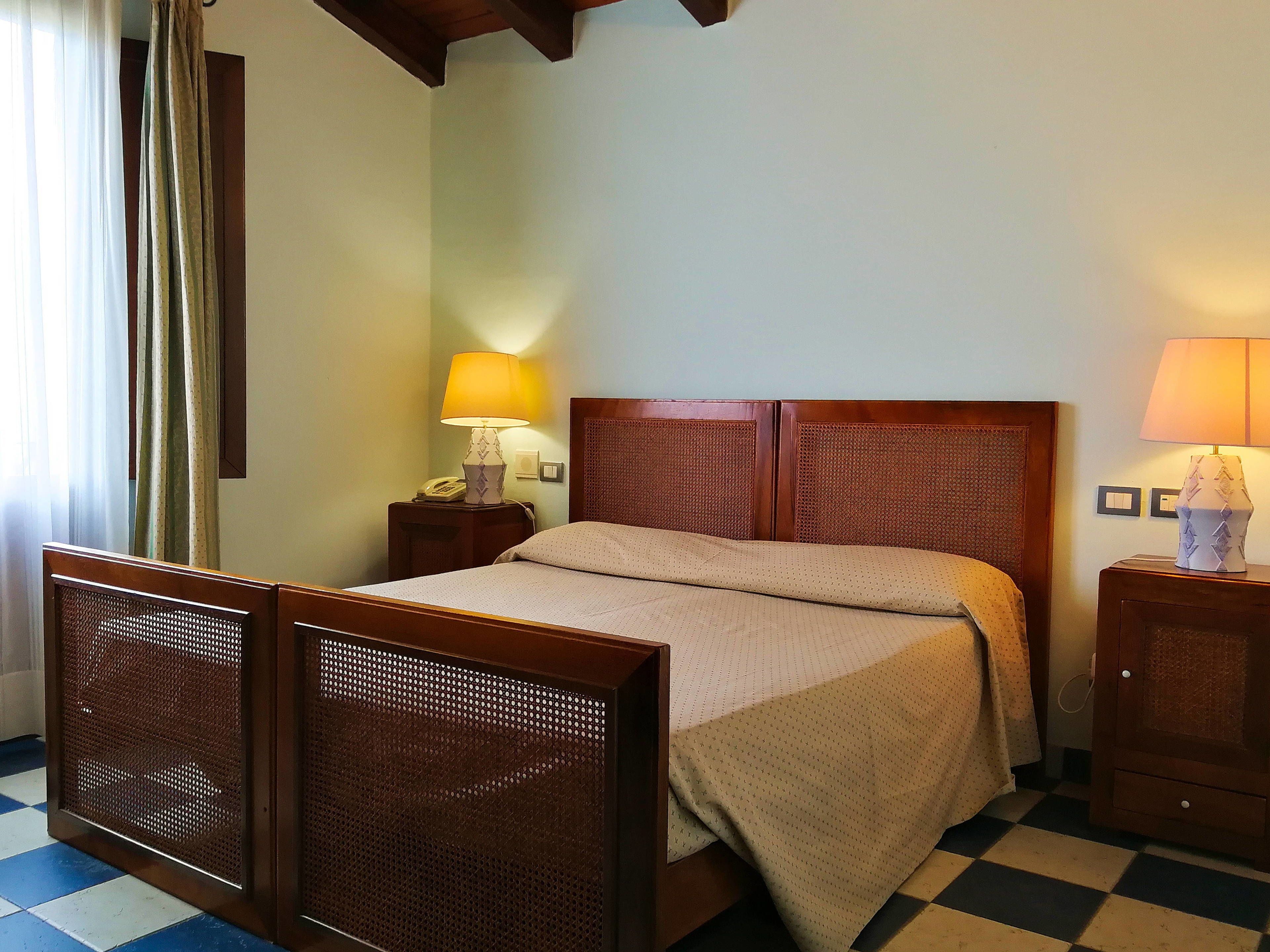 Habitación de hotel con piso a cuadros, cama de madera con cabecera, dos mesitas de noche con lámparas y cortina en la ventana.