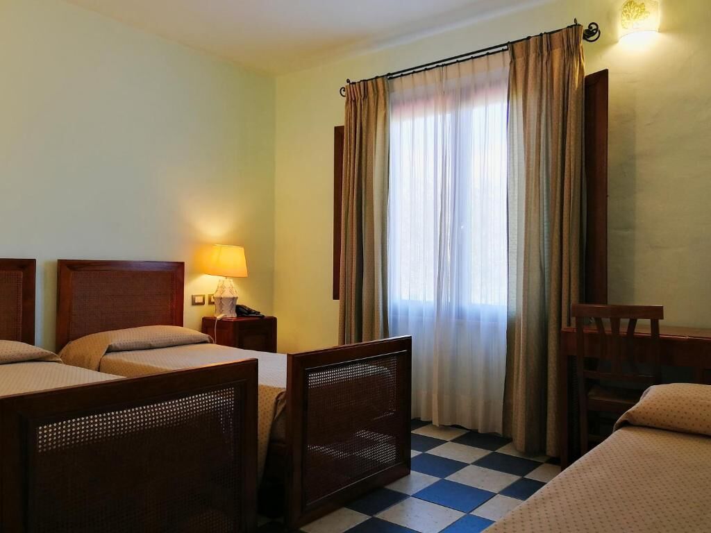 A hotel room with two beds, a chair, a nightstand with a lamp, and a window with curtains.