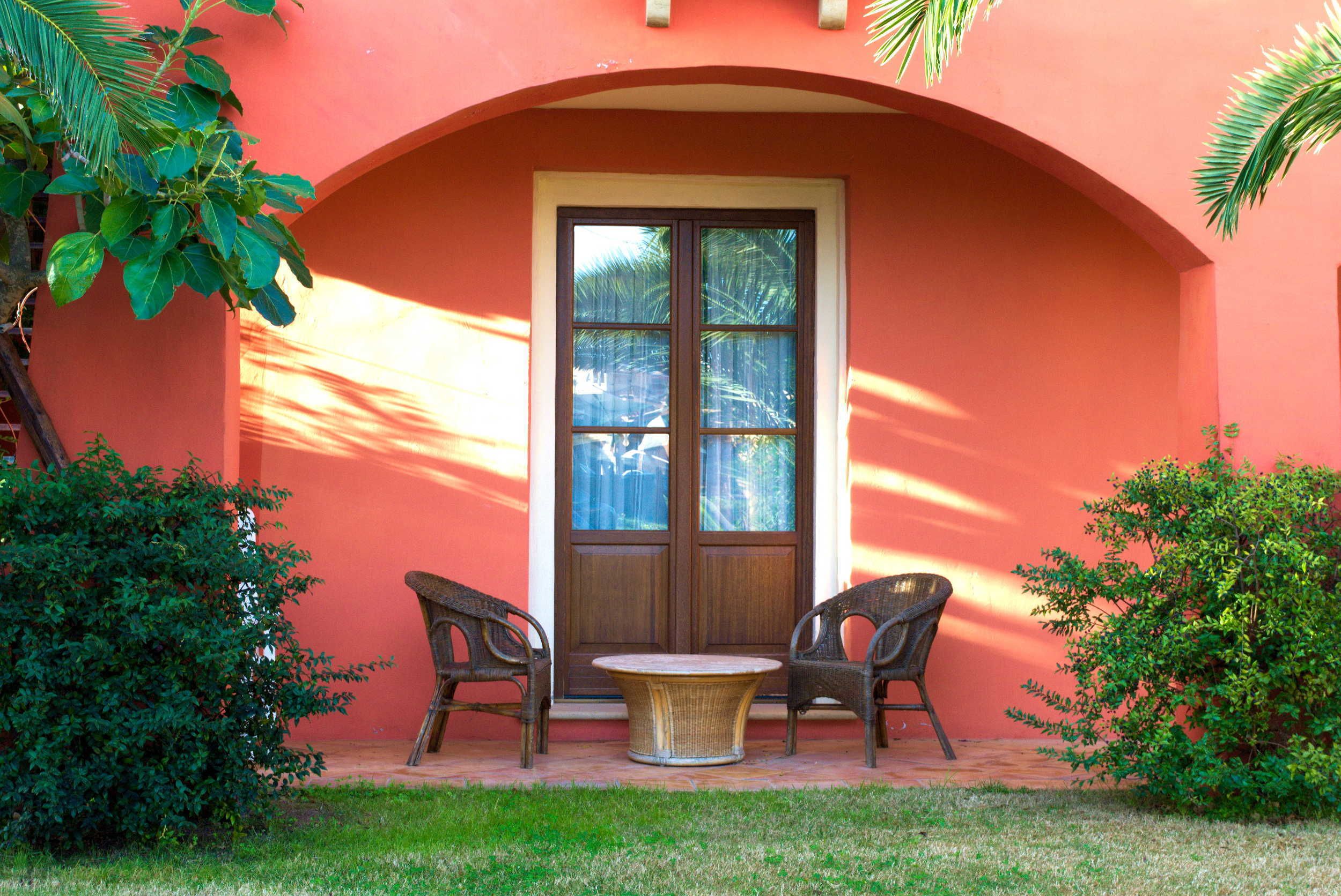 An outdoor seating area with two wicker chairs and a small round table, a large window, and a red wall.