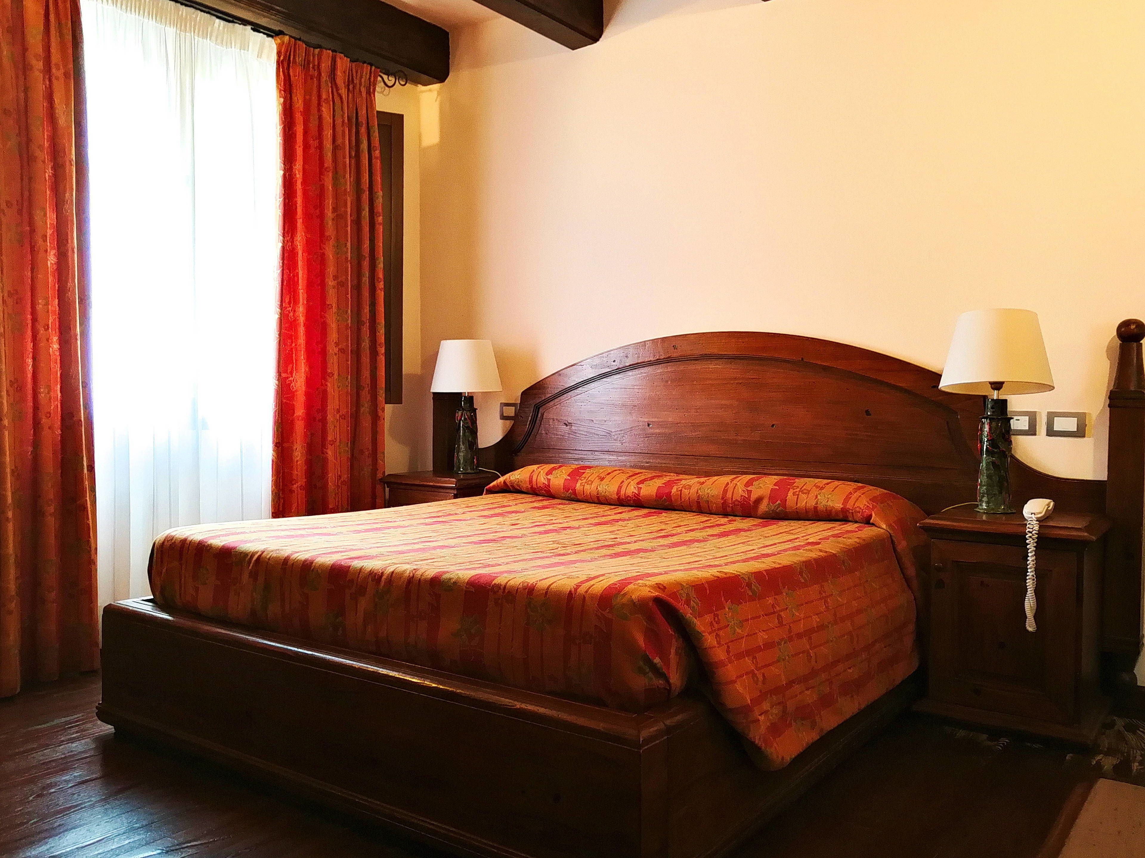 A hotel room with a wooden bed, two bedside tables with lamps, and a window with red curtains.