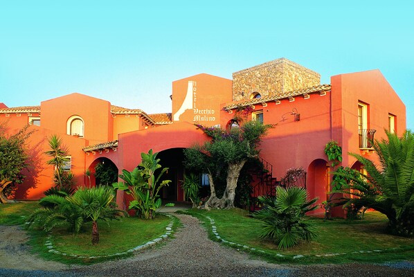 A hotel with a red facade, multiple arches, and a gravel pathway leading to the entrance. - Hotel Il Vecchio Mulino (Tortoli)