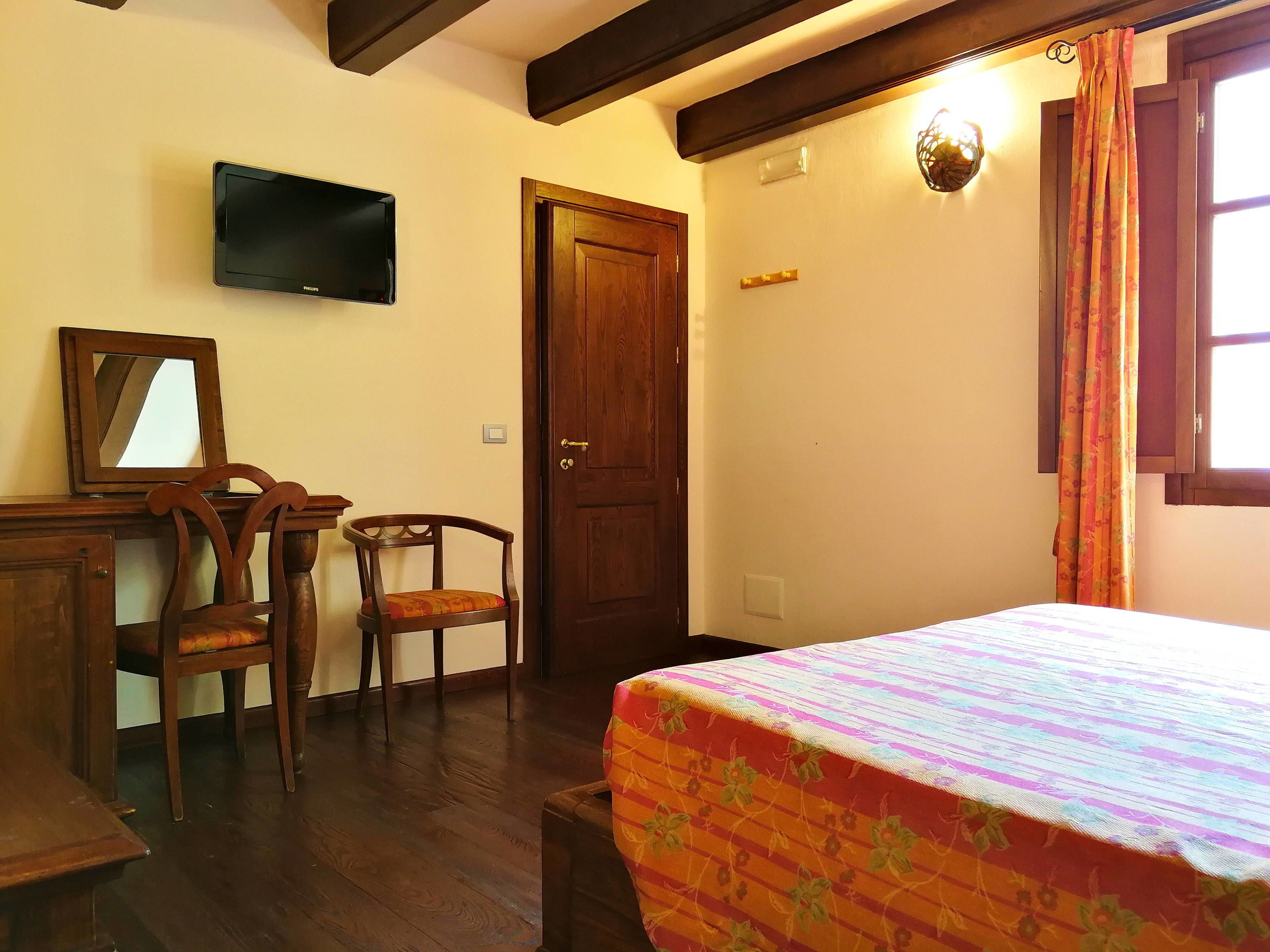 Una habitación con cama, un escritorio con silla, una puerta de madera, un televisor montado en la pared y una ventana con cortinas.