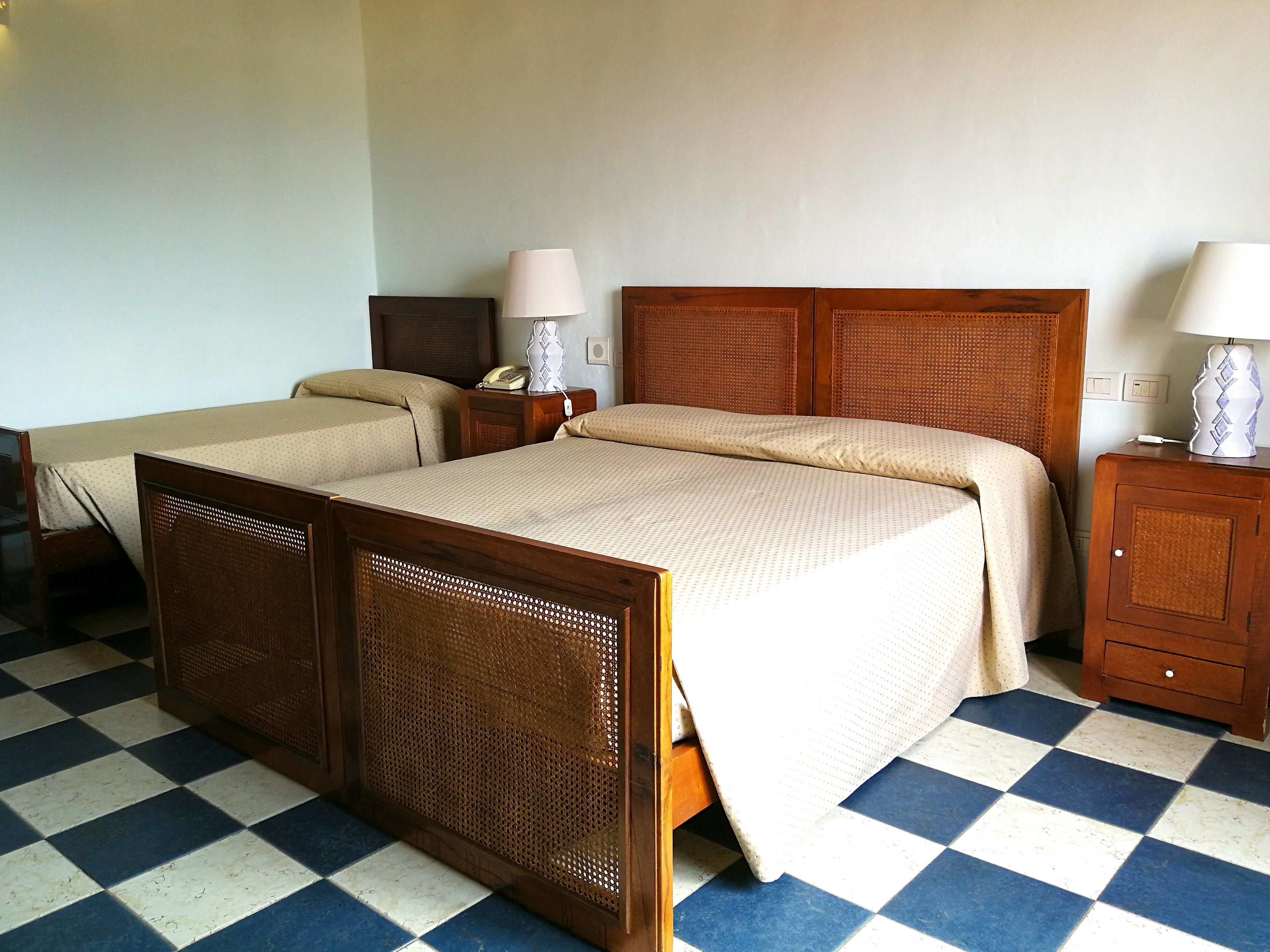 Une chambre d’hôtel avec un sol à carreaux, un lit en bois avec une tête de lit en osier, deux tables de chevet avec des lampes et une petite armoire en bois.