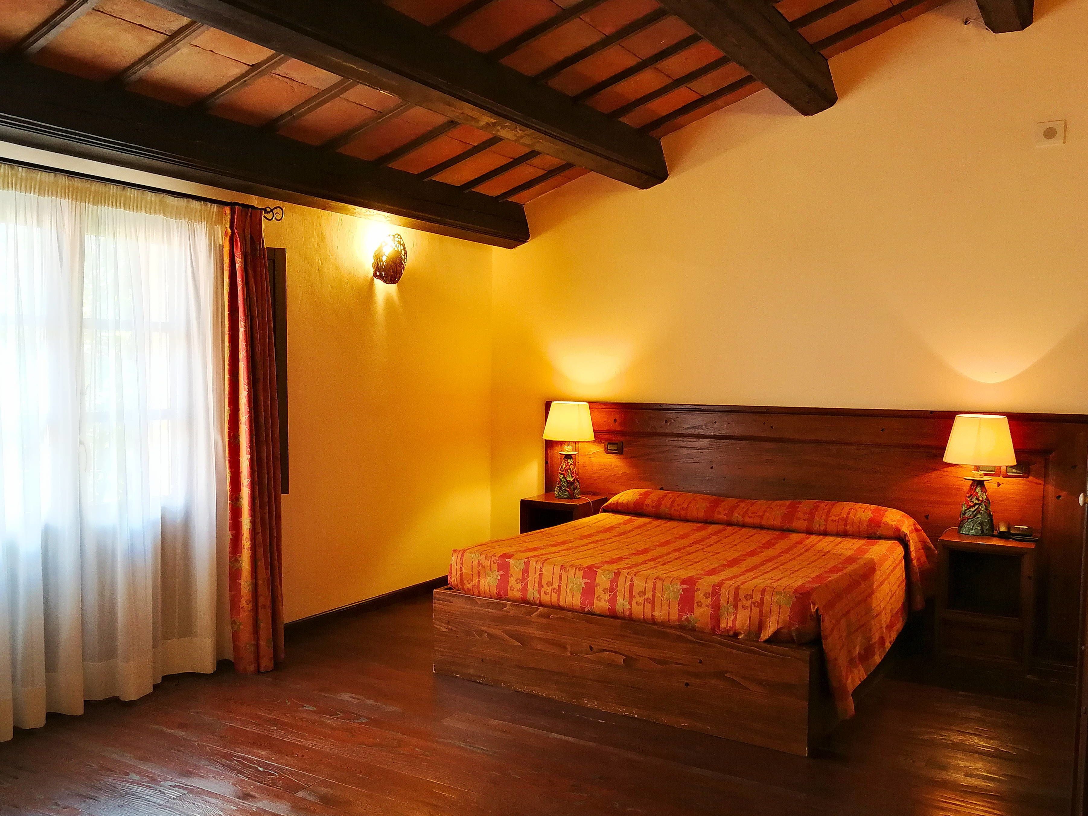 A hotel room with a wooden bed, two bedside tables with lamps, a wooden ceiling, and a window with sheer curtains.