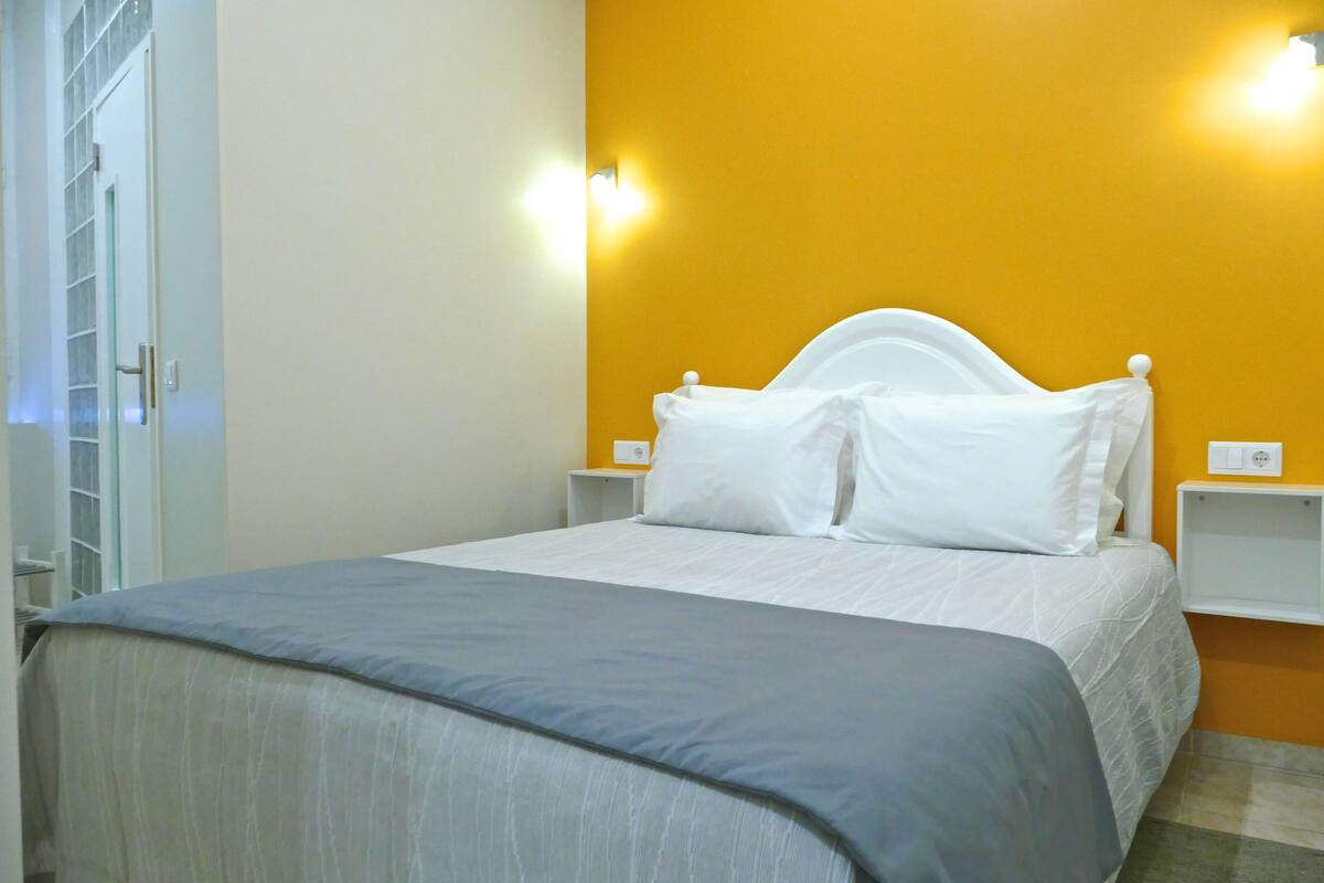 A neatly made bed with white linens in a room with a yellow accent wall and a white door.