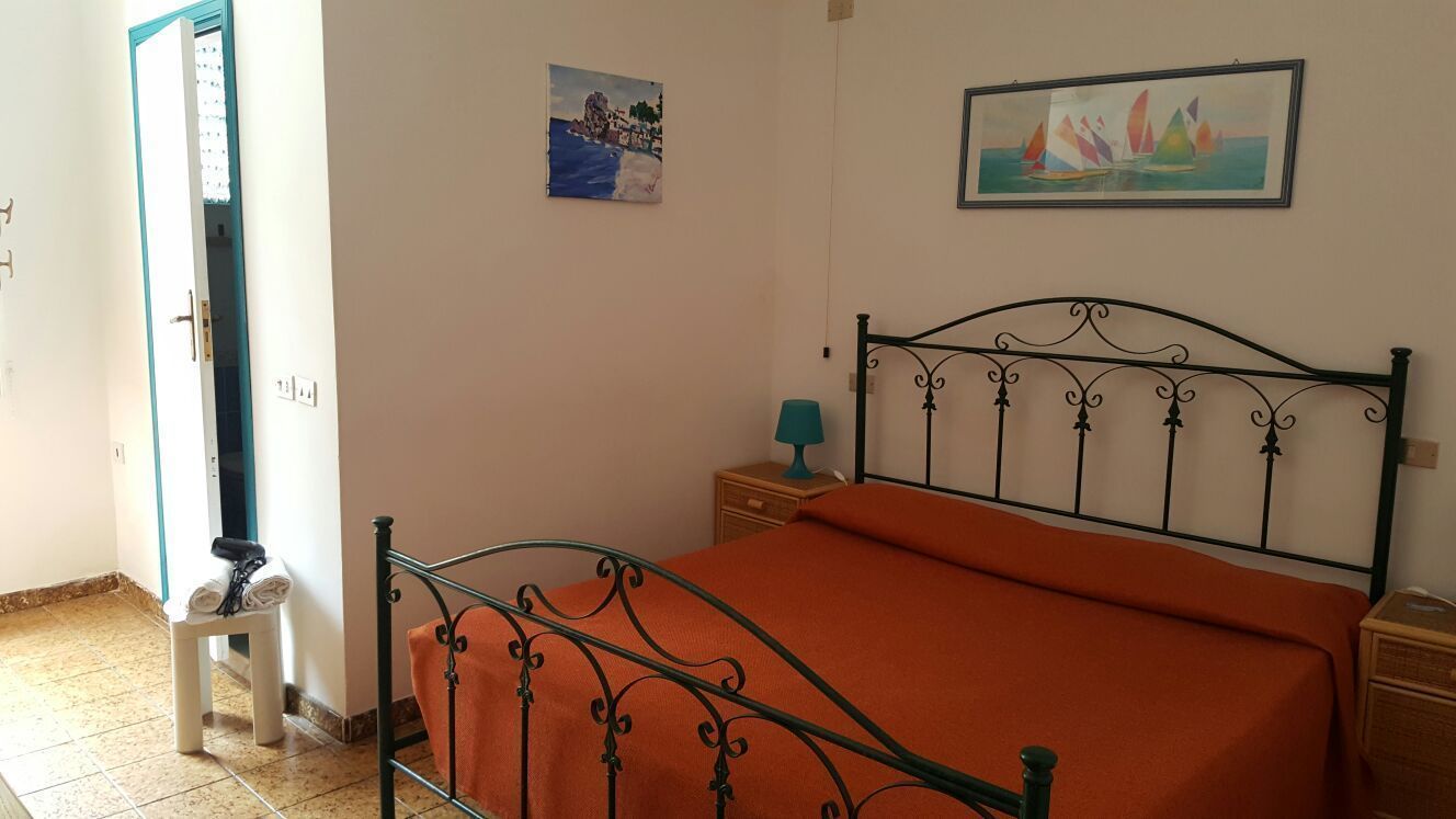 A bedroom with a metal bed frame, an orange bedspread, a small bedside table with a lamp, and two paintings on the wall.