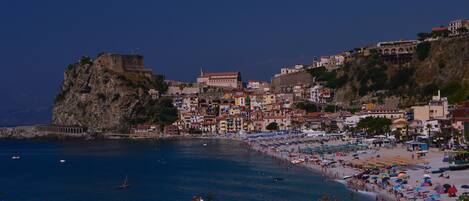 A coastal town with a historic fort on a cliff, a bustling beach, and colorful buildings.