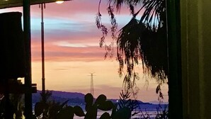A dining area with a large window overlooking a sunset, a table with chairs, and a hanging plant. - Le Sirene Scilla (Scilla)