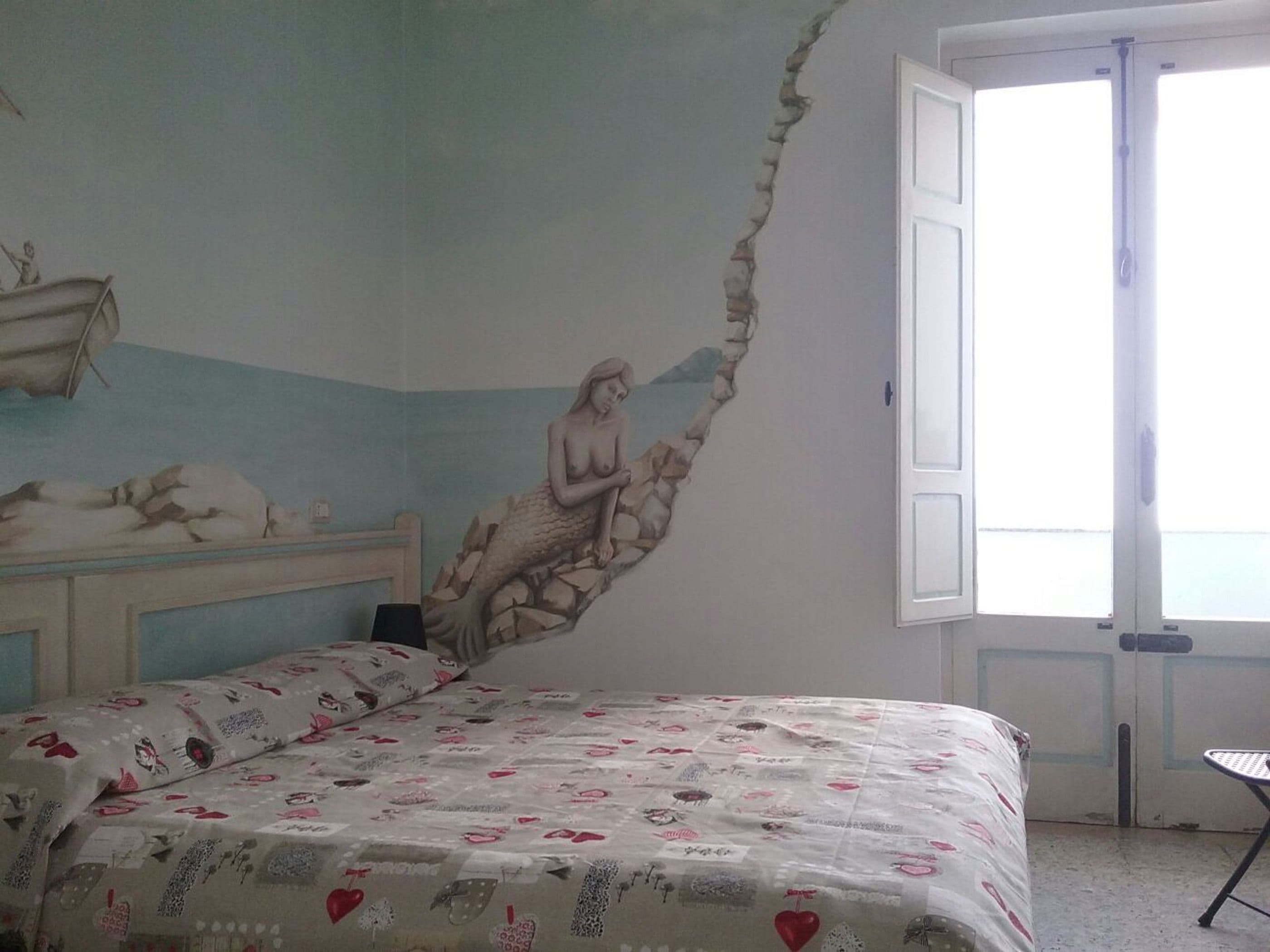 Een slaapkamer met een muurschildering van een zeemeermin, een bed met een geborduurde spreidek en een raam met uitzicht op zee.