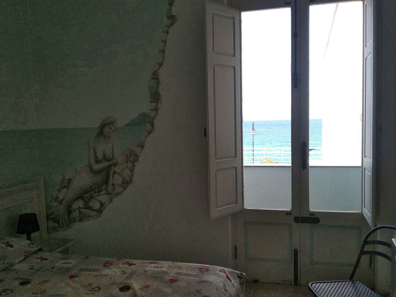 A bedroom with a mural on the wall, a bed with a floral bedspread, a chair, and a window with an open door revealing a view of the sea.