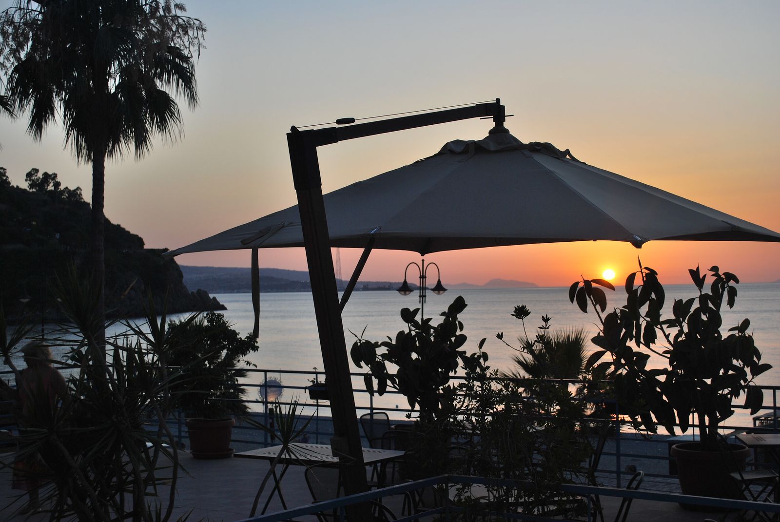 Ein Sonnenuntergangsblick von einer Terrasse mit Außenbestuhlung, mit Blick auf ein Gewässer und entferntes Land.