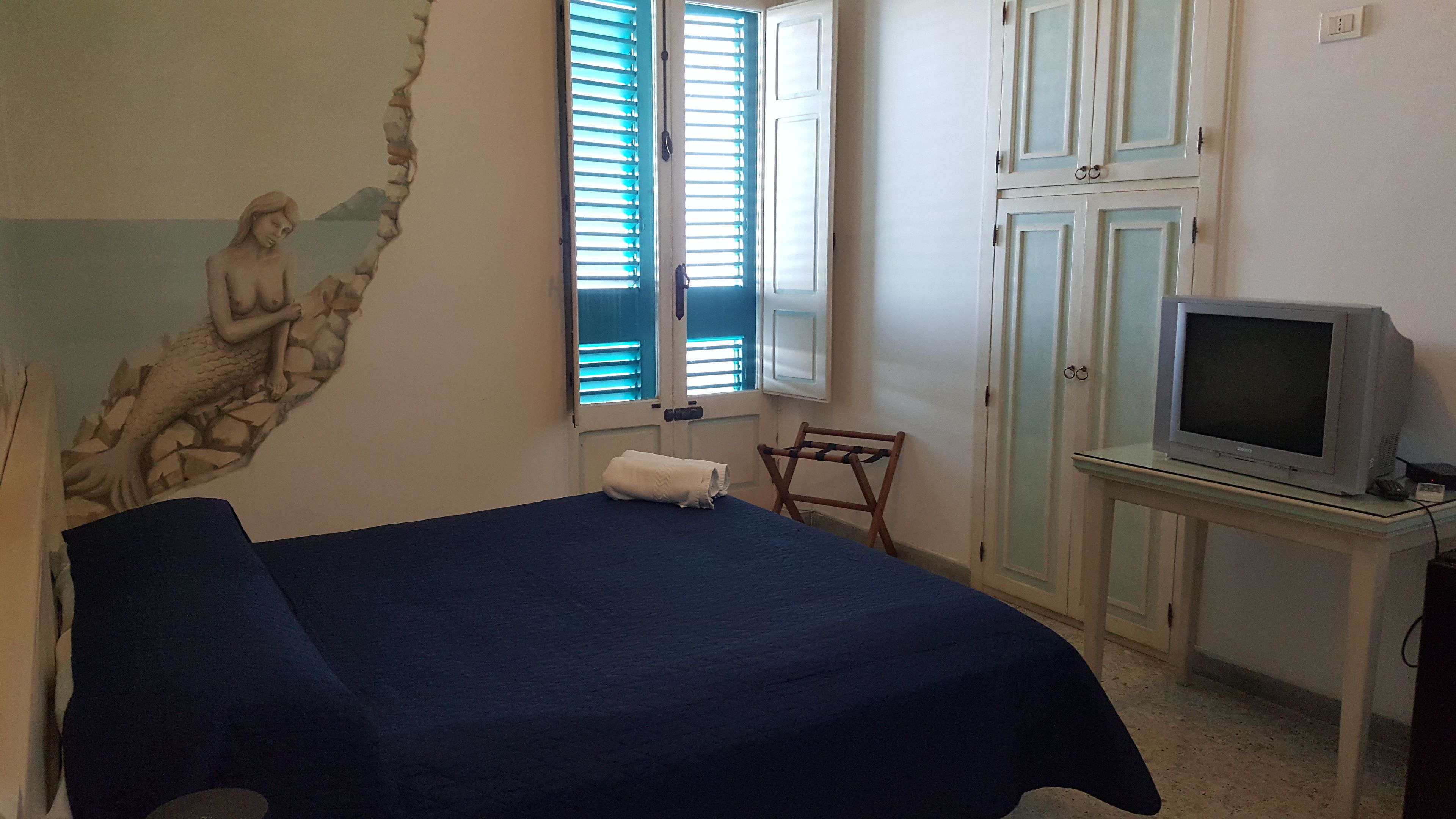 Ein Schlafzimmer mit einem Bett, einem Fernseher auf einem Schrank, einem Fenster mit Jalousien und einer Wand mit Wandgemälde.
