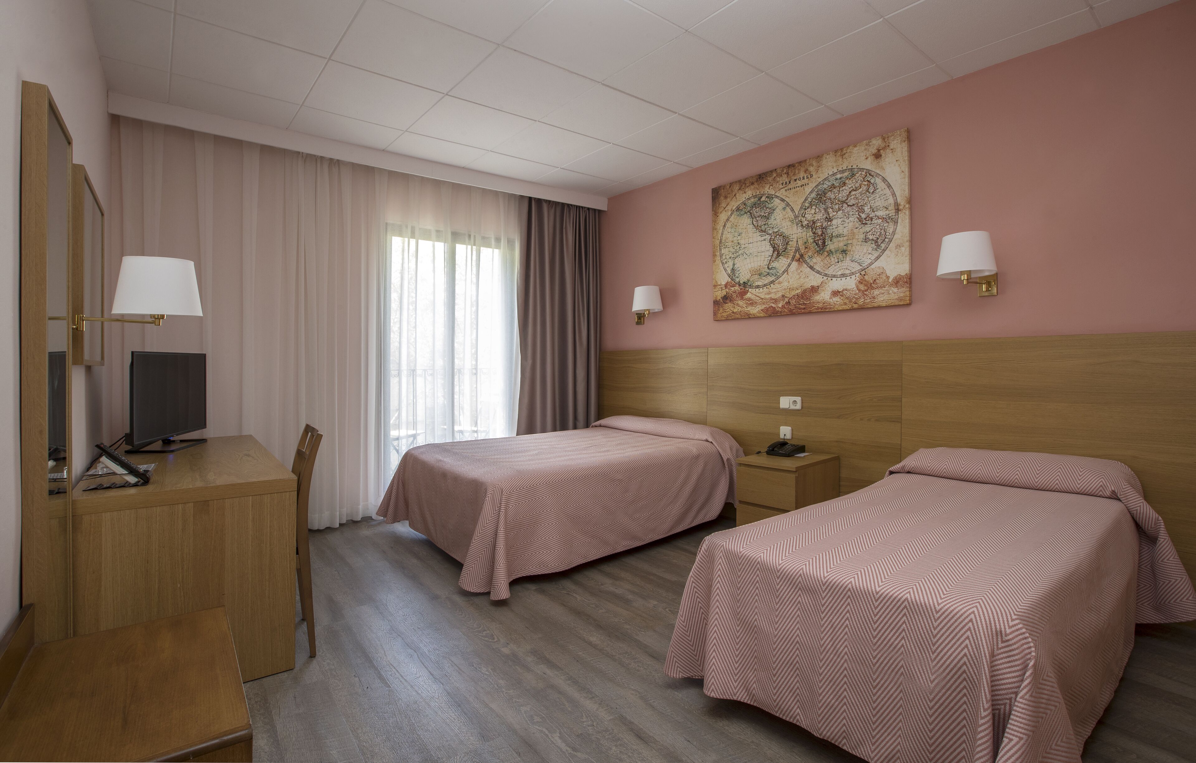 Habitación de hotel con dos camas individuales, un escritorio con ordenador, una silla y un ventanal con cortinas.