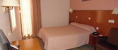 A hotel room with a bed, a desk with a computer, a chair, a nightstand with a lamp, and a painting on the wall.
