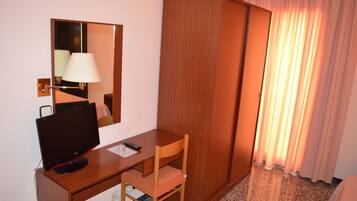 A hotel room with a wooden desk, a computer monitor, a chair, a lamp, a mirror, and a wardrobe.