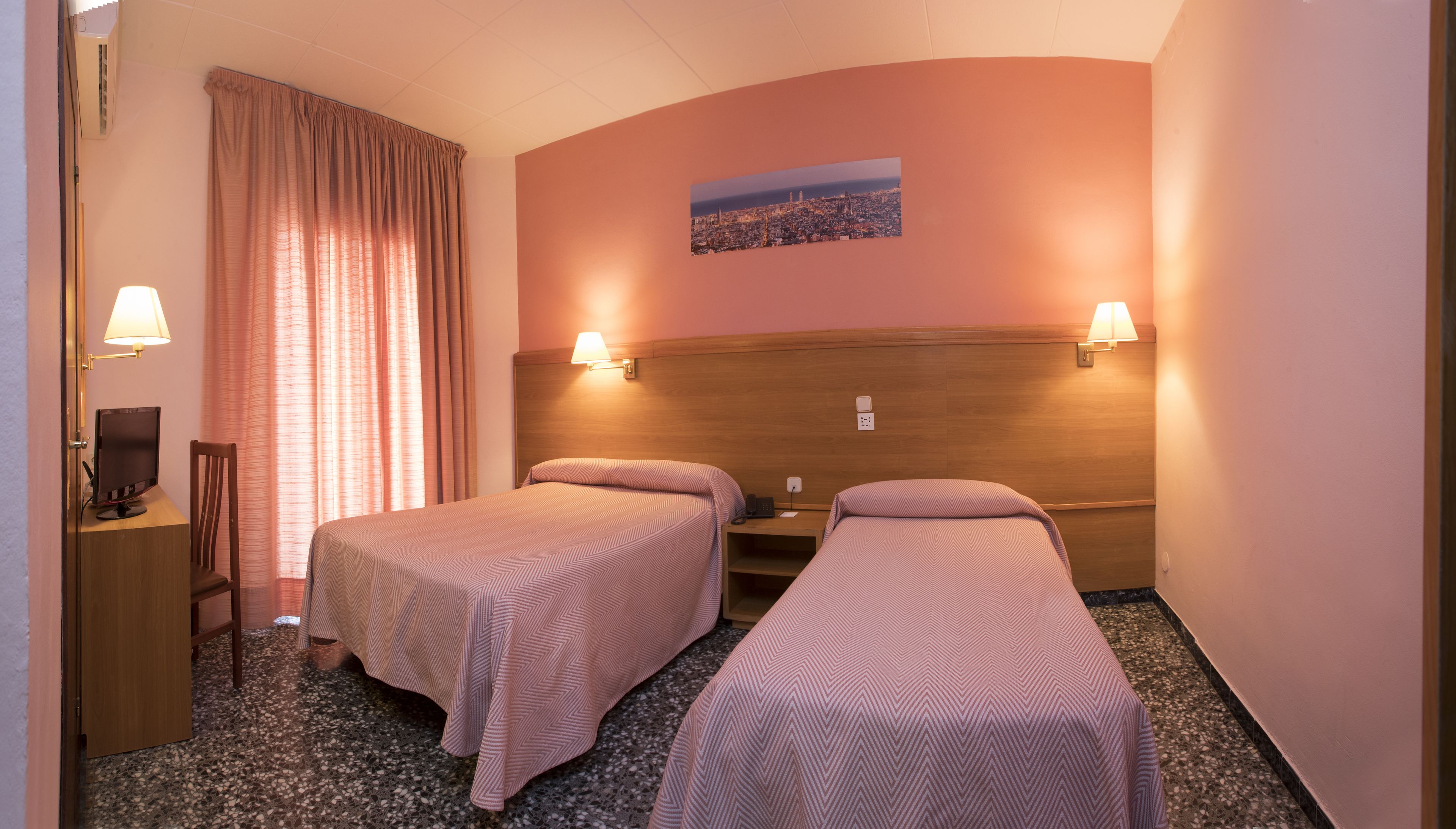 Une chambre d’hôtel avec deux lits simples, un bureau avec un ordinateur, une chaise, une fenêtre avec des rideaux et un tableau représentant une vue sur la ville.