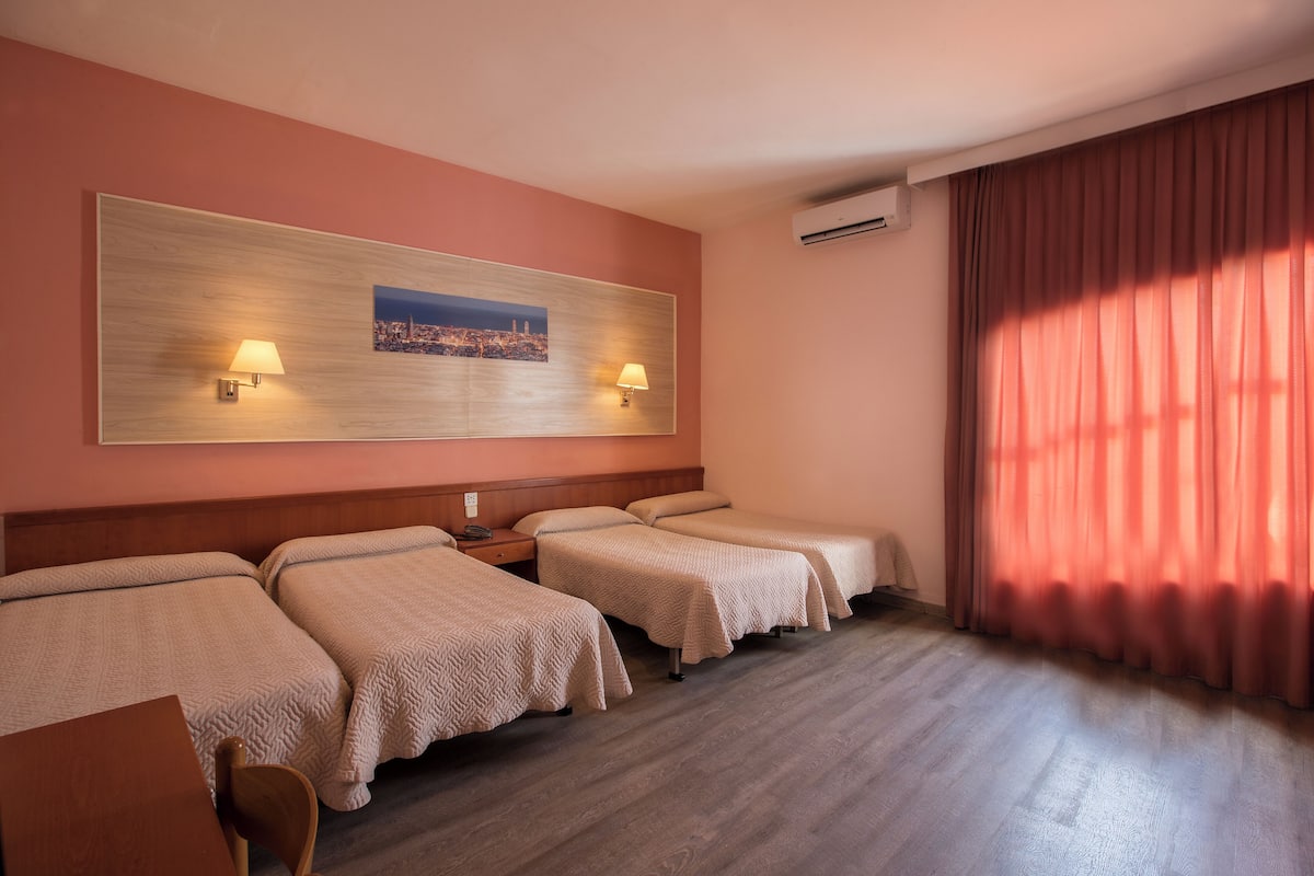 Une chambre d’hôtel avec trois lits, une tête de lit en bois, deux lampes fixées au mur, un climatiseur et une grande fenêtre avec des rideaux.