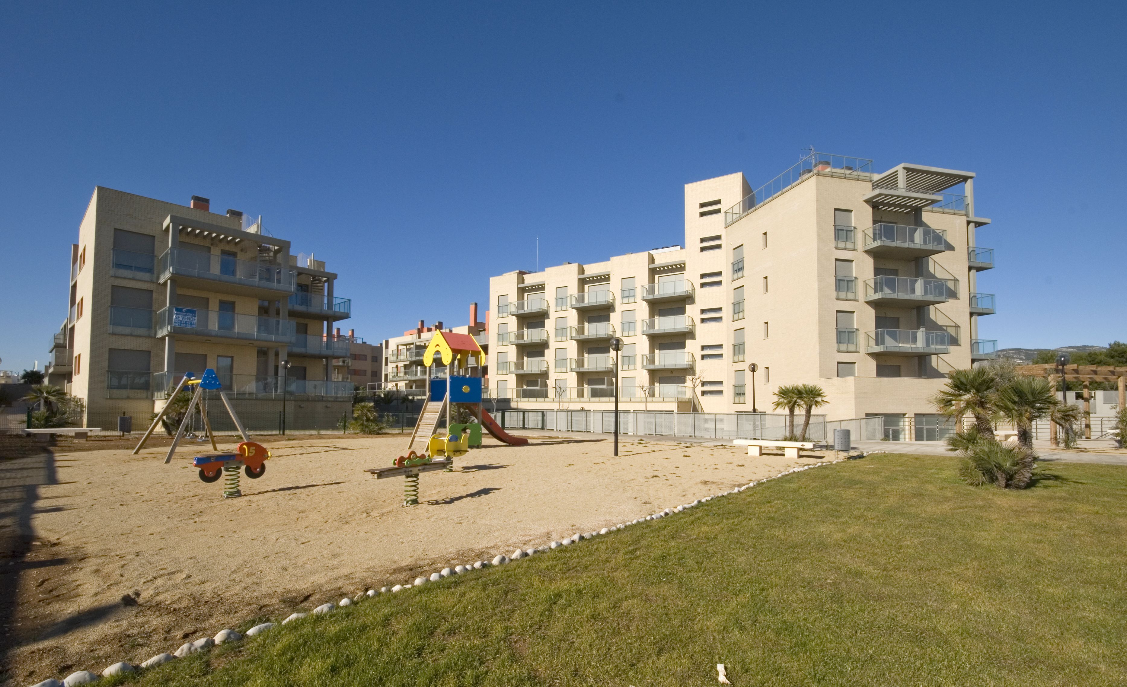 A playground with swings and slides, surrounded by a sandy area and grass. In the background, there are multi-story buildings with balconies.
