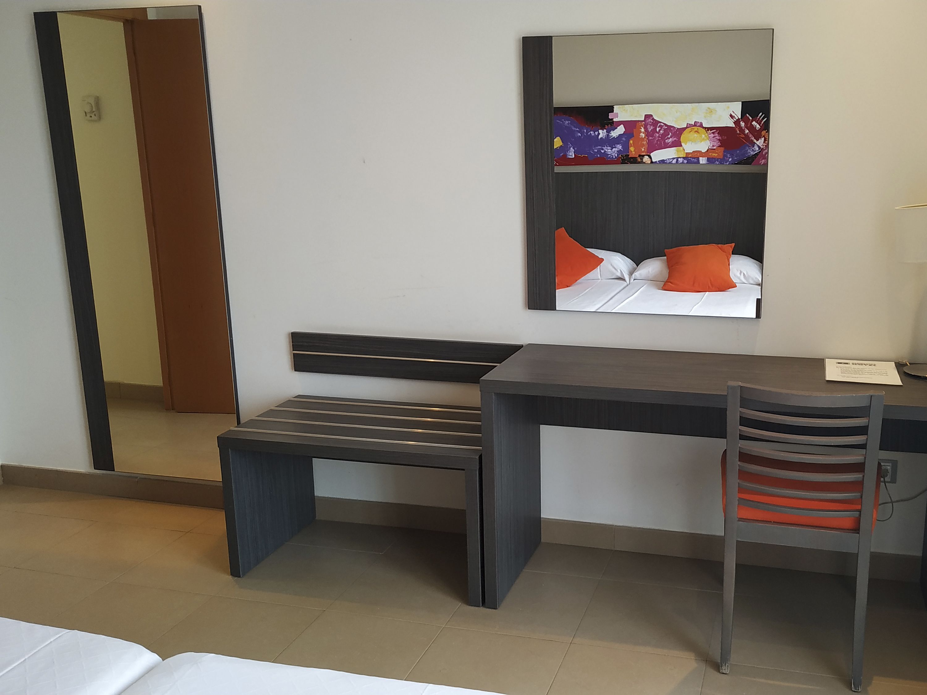 Une chambre d’hôtel équipée d’un lit, d’un bureau, d’une chaise, d’un miroir et d’une œuvre d’art accrochée au mur.