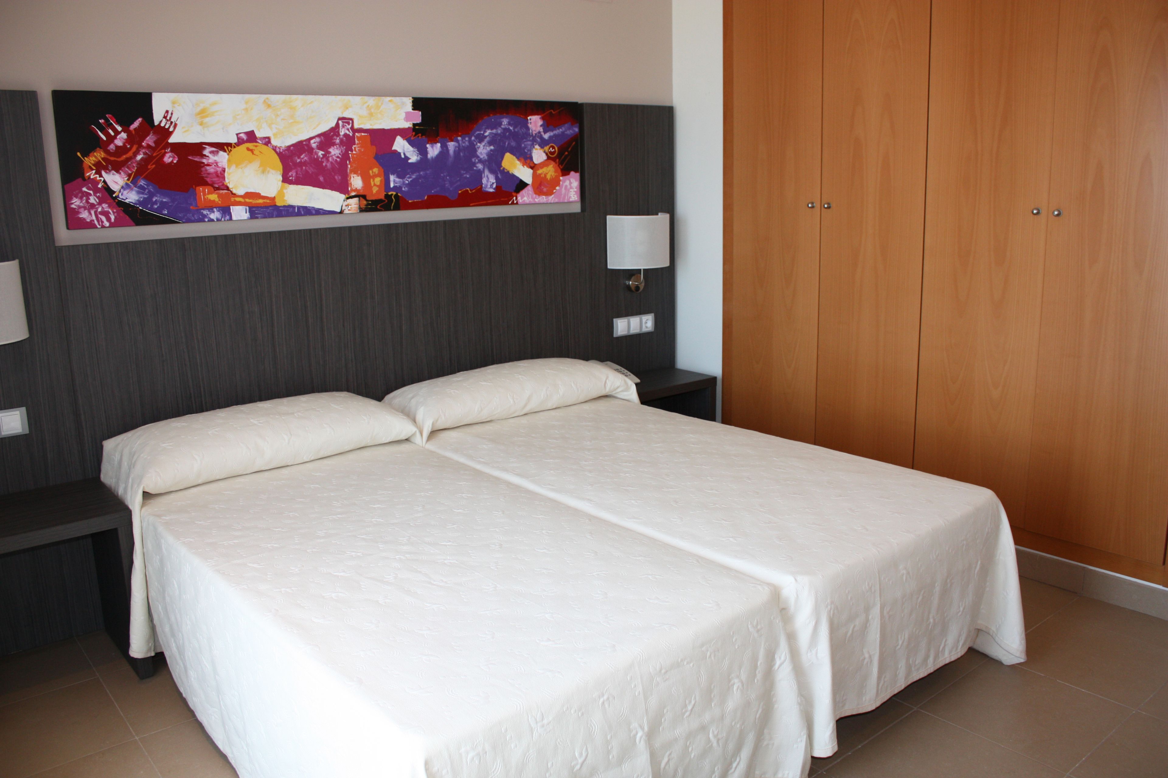 A double bed with a white bedspread, a nightstand with a lamp, a wooden wardrobe, and a colorful abstract painting on the wall.